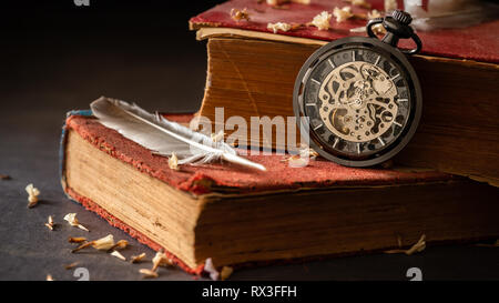 Winding pocket watch on old books with feathers and dried flower petals on the marble table in darkness and morning light.