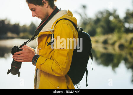 Close up of a man standing near a lake wearing a backpack looking at his dslr camera. Side view portrait of a tourist standing outdoors looking at his Stock Photo