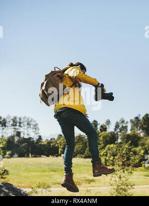 Explorer walking through tough terrain in the countryside holding a dslr camera. Rear view of a man wearing backpack jumping over a wooden log holding Stock Photo