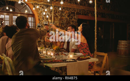 Group of friends making a toast with drinks at party. Young people sitting at a table with food, toasting drinks and enjoying dinner together. Stock Photo