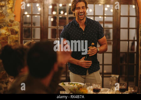 Laughing man opening champagne with friends sitting around table during party. Millennials having a dinner party. Stock Photo