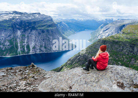 Girl in red jacket sits on rock and looks at mountains near Trolltunga. Popular tourist attraction. Ringedalsvatnet - lake in the municipality of Odda Stock Photo