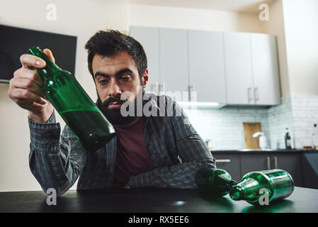 Drinking trouble. Depressed bearded man sits at the table with beer bottle in his hand. He is thinking about problems at work and troubles in relation Stock Photo