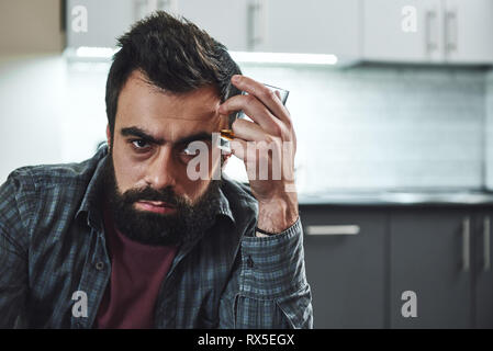 Drinking trouble. Depressed bearded man sits at the table with a glass of whiskey in his hand. He is thinking about problems at work and troubles in r Stock Photo