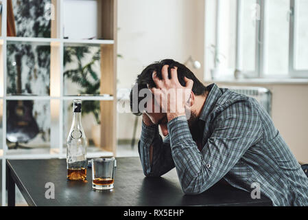 Drinking trouble. Depressed man sits at the table with his head in his hands. A bottle and a glass of whiskey stand in front of him. Side view Stock Photo