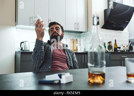 Dark-haired, sad and wasted alcoholic man sitting at the table, in the kitchen, after drinking whiskey and taking drugs, completely drunk, looking dep Stock Photo