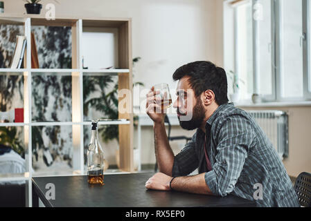 Drinking trouble. Depressed bearded man sits at the table with a glass of whiskey in his hand. He is thinking about problems at work and troubles in r Stock Photo