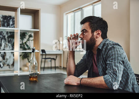 Drinking trouble. Depressed bearded man sits at the table with a glass of whiskey in his hand. He is thinking about problems at work and troubles in r Stock Photo