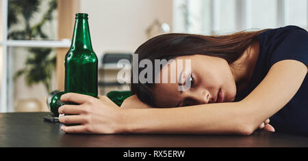 Dark-haired, sad and wasted alcoholic woman lying on a table, in the kitchen, drinking beer, holding bottle, completely drunk, looking depressed, lone Stock Photo