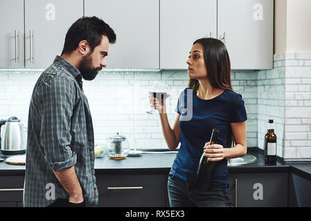 Drunkard. Dark-haired addicted woman arguing with her husband. She is standing in the kitchen with a glass and a bottle of red wine and listening to h Stock Photo