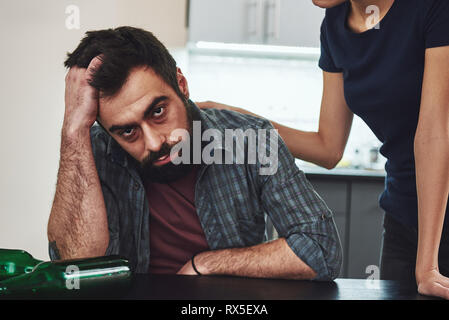 Drunkard. Dark-haired addicted woman feeling despair while standing near her drunk husband. He is sitting in the kitchen, wasted. Empty bottles are on Stock Photo