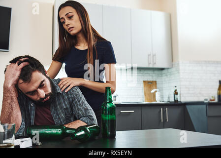 Drunk husband. Dark-haired addicted woman feeling despair while standing near her drunk husband. He is sitting in the kitchen, wasted. Empty bottles a Stock Photo