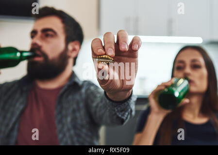 Young woman and her man are standing in the kitchen, drinking beer from bottles. Man shows bottle cap. They both have depression, both suffer from alc Stock Photo