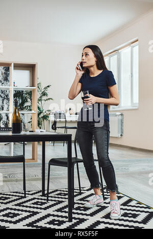 Dark-haired, sad and wasted alcoholic woman standing in the kitchen, holding a glass of wine, while talking on the phone at home. She is completely dr Stock Photo