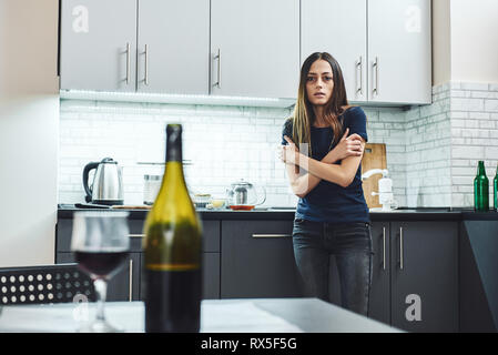 Dark-haired, sad and wasted alcoholic woman standing in the kitchen, looking at red wine on the table, completely drunk, looking depressed, lonely and Stock Photo