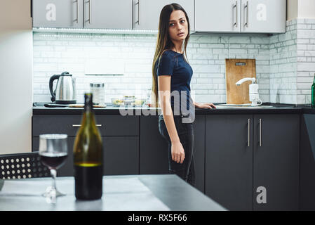 Dark-haired, sad and wasted alcoholic woman standing in the kitchen, looking at red wine on the table, completely drunk, looking depressed, lonely and Stock Photo