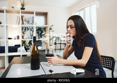 Portrait of young woman in despair, smoking cigarette while sitting at table, holding glass of alcohol at home. Female alcoholism concept. Protest in  Stock Photo
