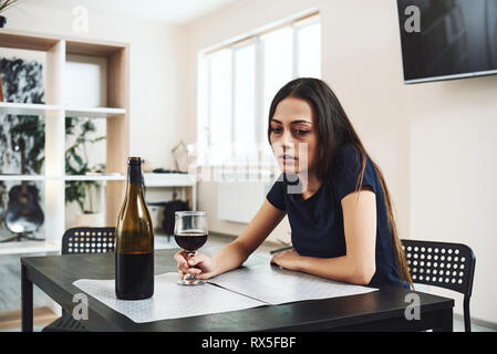 Dark-haired, sad and wasted alcoholic woman sitting at home, in the kitchen, drinking red wine, holding glass, completely drunk, looking depressed, lo Stock Photo