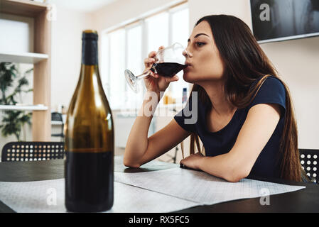 Depressed, divorced woman sitting alone in kitchen at home and drinking a glass of red wine because of problems at work and troubles in relationships. Stock Photo