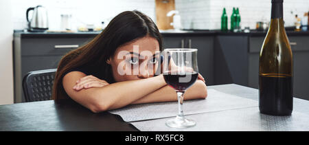 Dark-haired, sad and wasted alcoholic woman sitting at home, in the kitchen, leaning over the table, looking at bottle and glass of red wine on the ta Stock Photo