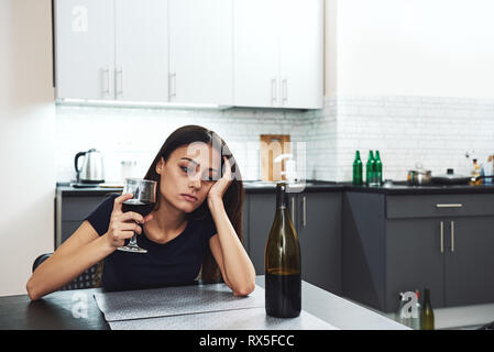 Dark-haired, sad and wasted alcoholic woman sitting at home, in the kitchen, leaning over the table, holding a glass and looking at bottle of red wine Stock Photo