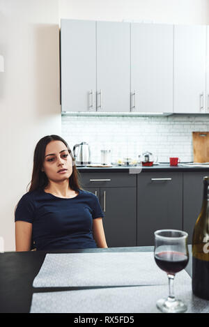 Dark-haired, sad and wasted alcoholic woman sitting at home, in the kitchen, looking at bottle and glass of red wine on the table, completely drunk, l Stock Photo