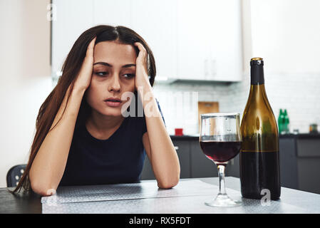 Dark-haired, sad and wasted alcoholic woman sitting at home, in the kitchen, leaning over the table, looking at bottle and glass of red wine on the ta Stock Photo