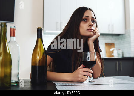 Dark-haired, sad and wasted alcoholic woman sitting at home, in the kitchen, drinking red wine, holding glass, completely drunk, looking depressed, lo Stock Photo