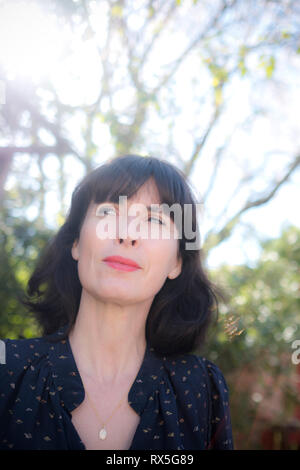 close up of beautiful french woman thinking to herself with a sideways glance and a smile with light up trees and garden in background Stock Photo