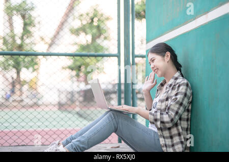 Hi on video chat. Cute teenage having a video call with a laptop greeting a friend lying on green grass loan in a campus park on background Stock Photo