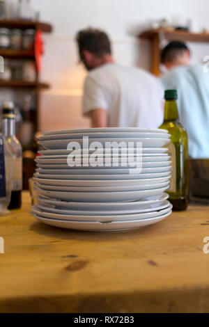 A stack of chipped white plates sit on a wooden pine table in foreground in kitchen with two men cooking in background Stock Photo