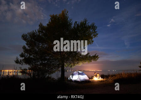 Tourist tent under big tree and young couple, bearded man and pretty woman hikers having a rest at night on sea shore by campfire under blue evening s Stock Photo