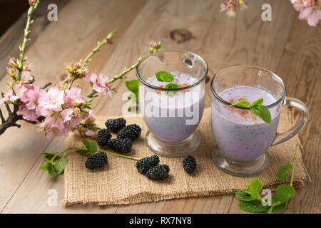 Fresh fruit smoothie blackberry milkshake, purple yogurt or ice cream drink on wood table with sprigs of mint, blackberries, and cherry blossoms. Stock Photo