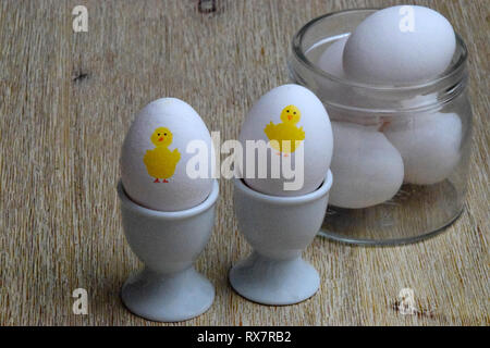 Two boiled eggs in egg cups with yellow Easter chicken painted on them and eggs in a glass bowl Stock Photo
