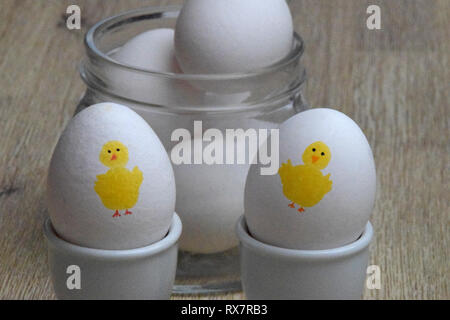 Boiled eggs with yellow Easter chicken painted on them in egg cups and with white eggs in a glass jar in the background Stock Photo