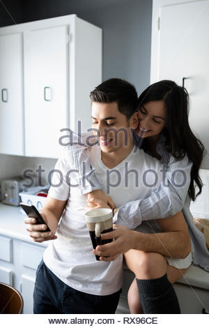 Affectionate young Latinx couple using smart phone and drinking coffee in kitchen
