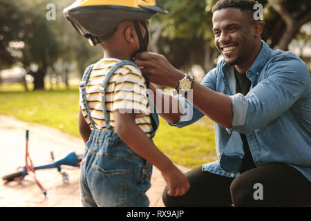 Smiling man helping son wearing helmet for cycling at park. Boy getting ready by wearing bike helmet to start cycling. Stock Photo