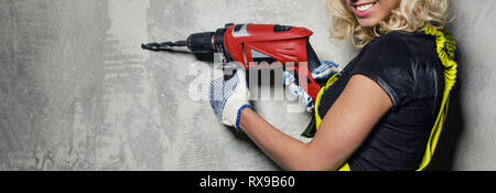 Attractive builder woman wearing protective coverall workwear smile look at camera holding a drill in her hands pose over obsolete horizontal wall Stock Photo