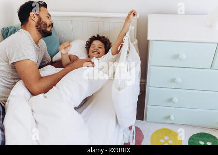 Man waking up his son lying on bed. Father lying beside his son on bed and talking to him. Stock Photo