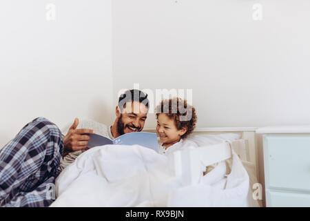 Happy father and son reading a book sitting on bed. Smiling boy enjoying reading a story with his father. Stock Photo