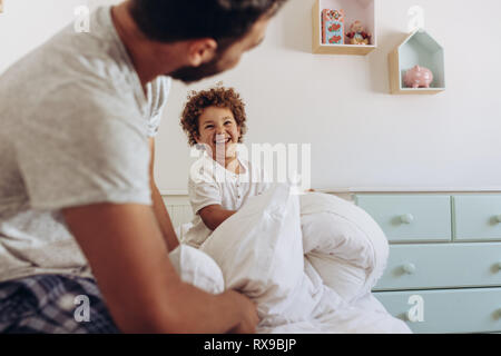Father and son having a pillow fight sitting on bed. Cheerful man and kid playing with pillows on bed. Stock Photo