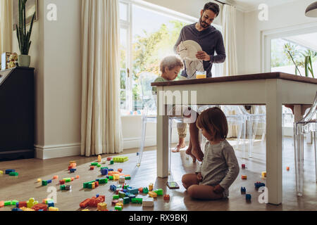 Man looking after his kids while doing the household works. Kid playing with toys sitting on floor with his father doing daily household chores. Stock Photo