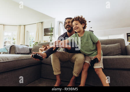 Father and son having fun playing video game at home. Man playing video game with son sitting on couch at home holding joysticks. Stock Photo