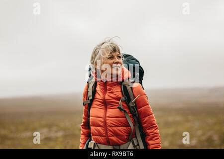 Senior woman standing on a hill wearing a backpack and looking away. Adventure seeking woman trekking on a hill. Stock Photo