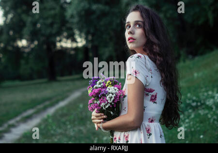 Side view of thoughtful woman holding colorful flowers while standing on grassy field at park Stock Photo