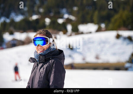 Portrait of smiling woman wearing ski goggles while standing on snow covered field Stock Photo