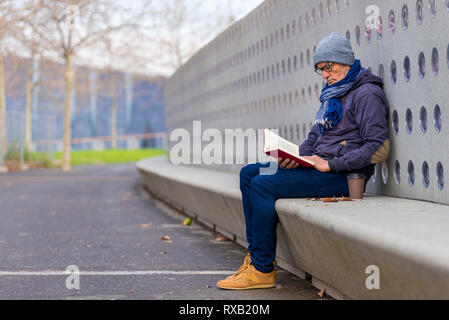 Fashionable senior man wearing warm clothing reading book on seat in park during autumn
