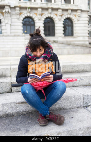 Full length of female tourist wearing warm clothing reading guidebook while sitting on steps in city Stock Photo