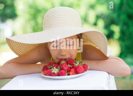 Portrait of Cute preteen girl in big hat is eating strawberries at summer day Stock Photo