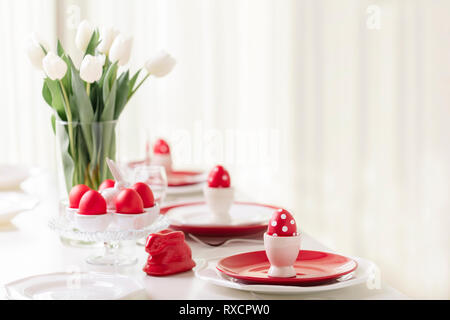 Happy easter. Decor and table setting of the Easter table is a vase with white tulips and dishes of red and white color. Easter colored eggs with whit Stock Photo